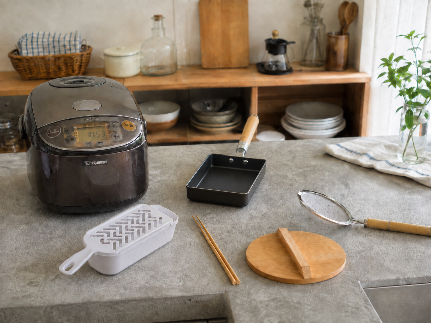 Japanese kitchen tools on a counter: a rice cooker, tamagoyaki pan, grater, cooking chopsticks, and a strainer